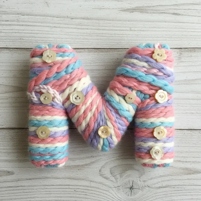 A cardboard letter M wrapped tightly with thick purple yarn and decorated with small white button dots.