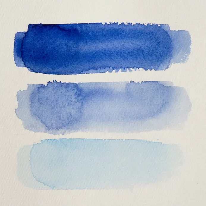 A brush loaded with watercolor paint creates three test strokes of different consistencies on a white palette.