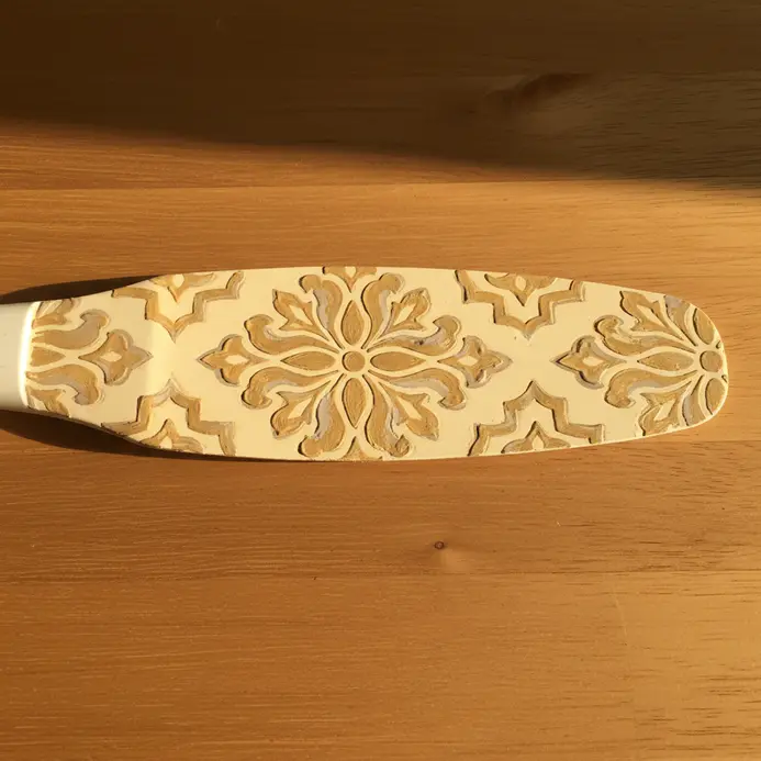 A cream-colored fan blade with a detailed, symmetrical damask pattern stenciled in gold paint.