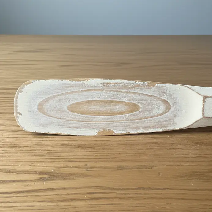 A wooden fan blade with layers of white paint lightly sanded to reveal the grain underneath.
