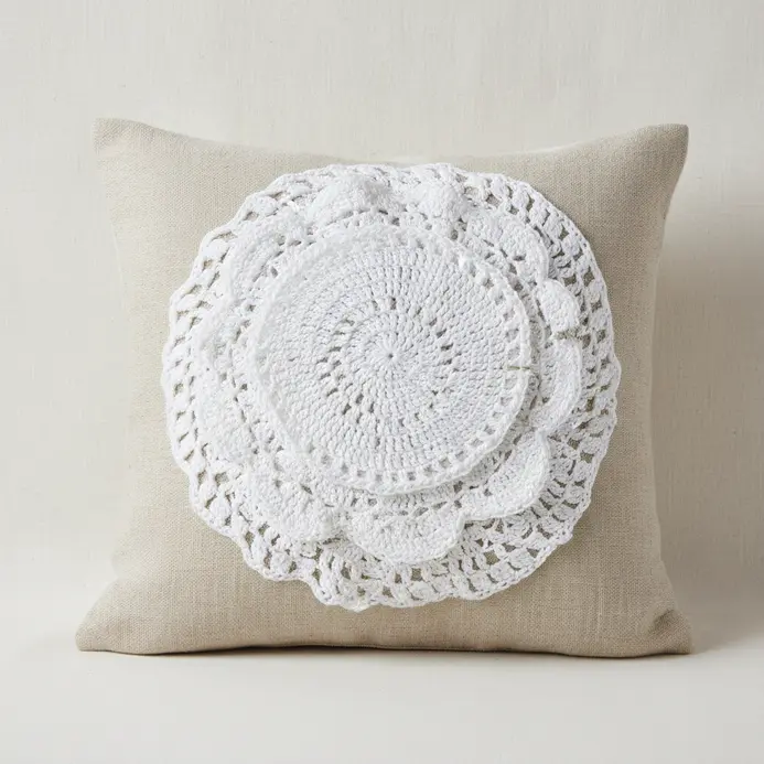 A neutral linen pillow cover with several overlapping white lace doilies sewn to the center.