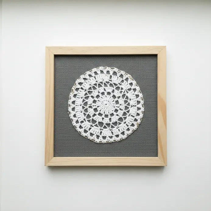 A white crocheted doily mounted against dark fabric inside a wooden shadow box frame.