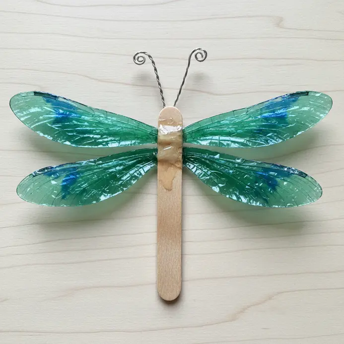 A dragonfly sculpture made from a popsicle stick body, with cellophane wings and curled pipe cleaner antennae.