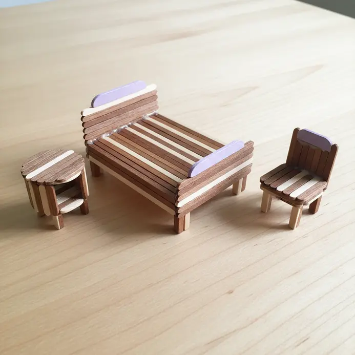 A small-scale bedroom furniture set including a bed, nightstand, and chair, meticulously built from popsicle sticks.