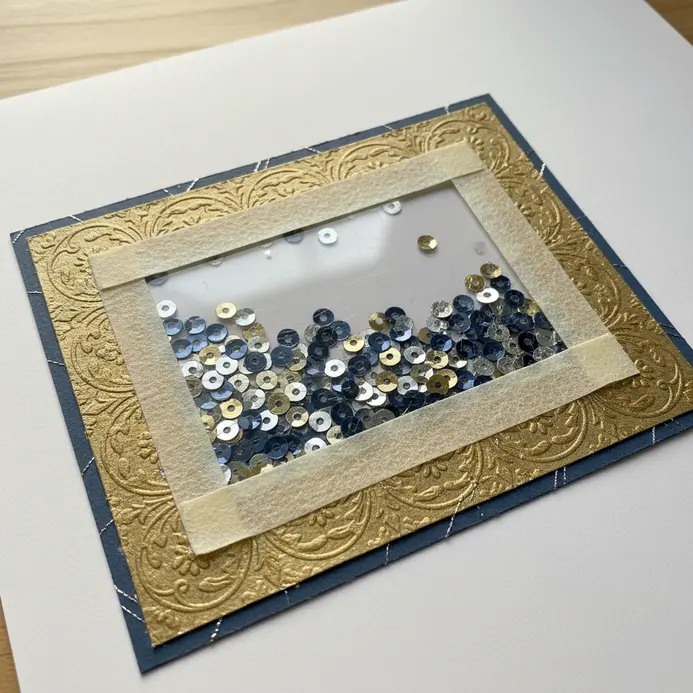 An interactive shaker box element on a scrapbook page, filled with gold and silver sequins behind a plastic window.