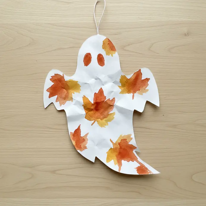 A white paper ghost cutout with a painted autumn leaf pattern, hanging from a string.