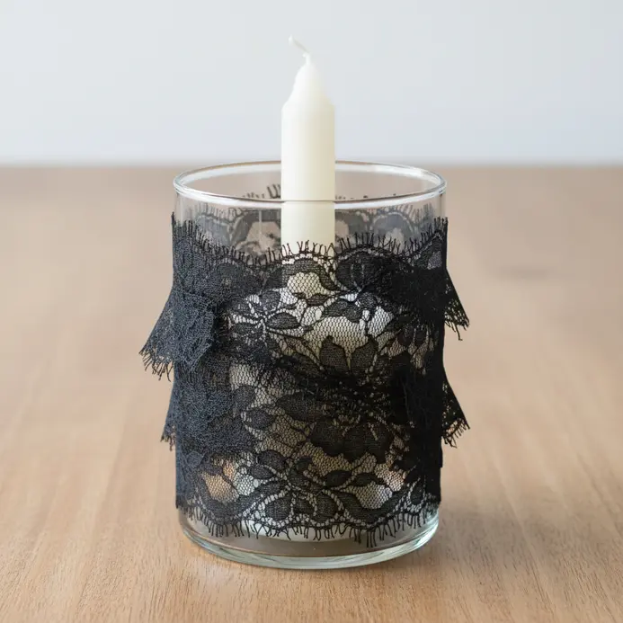 A clear glass votive candle holder wrapped with a band of delicate black lace, with a candle inside.