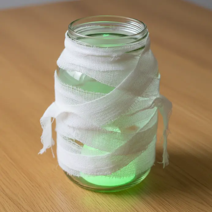 A glass mason jar wrapped in strips of white gauze, with a green glow stick illuminating it from within.