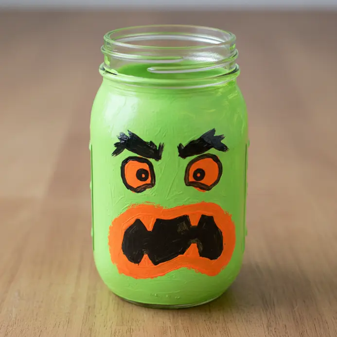 A glass mason jar painted with a bright green base and a playful neon orange monster face.