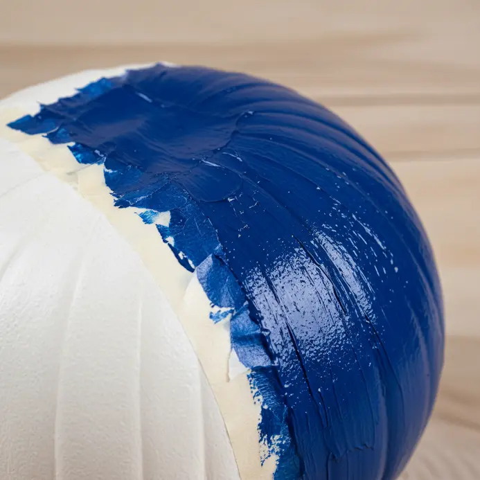 A brush applies a thick, even coat of cobalt blue paint to a taped-off pumpkin section.