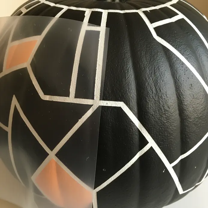 A hexagonal pattern stencil is pressed against a pumpkin, ready for paint application.