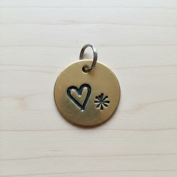 A circular brass keychain tag featuring a hand-stamped initial and a small heart design, with a jump ring attached.