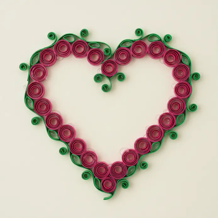 A piece of framed paper art where a heart shape is created from intricate quilled paper roses and green scrolls.