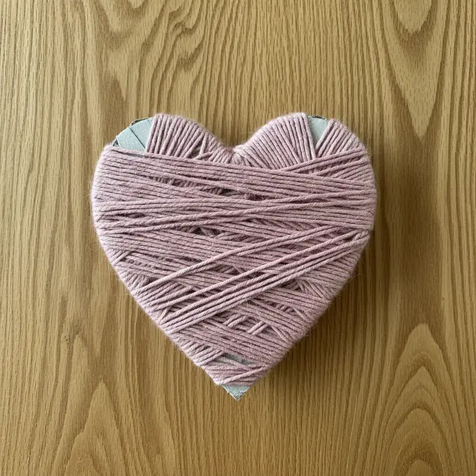 A thick cardboard heart form neatly wrapped in a continuous strand of soft pink yarn, creating a textured surface.
