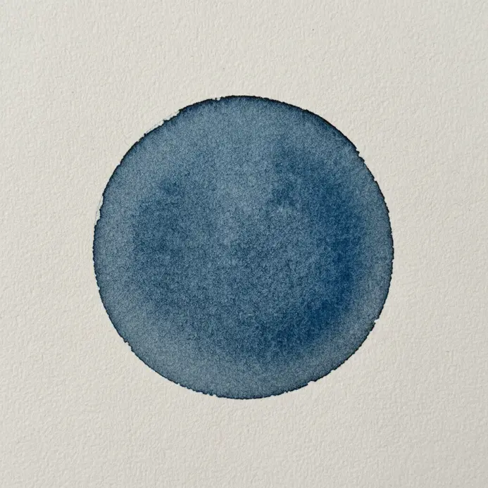 A perfectly round, dark blue droplet of watercolor paint on dry paper, with a slightly darker edge.