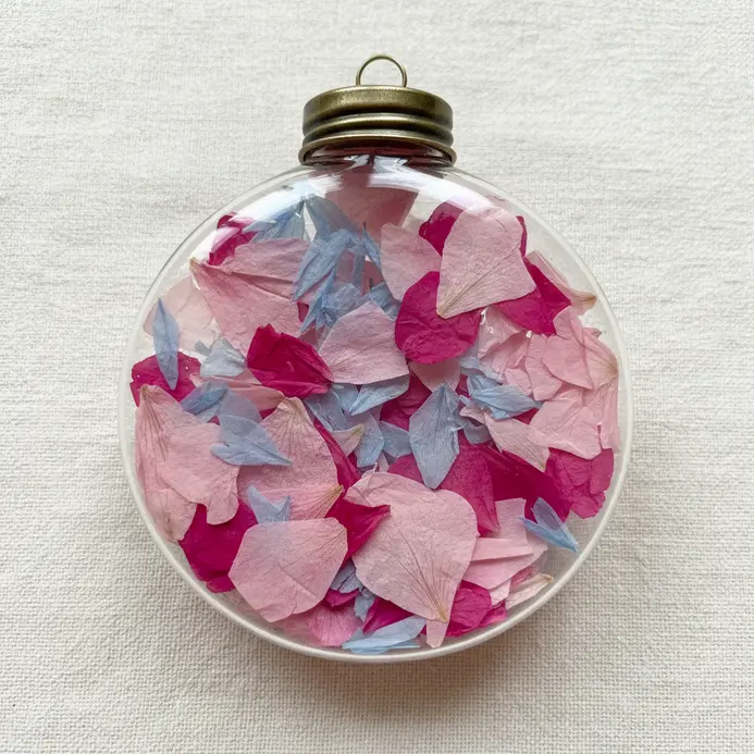 A clear glass ball ornament hanging from a ribbon, filled with tiny pieces of colorful pressed flower petals.