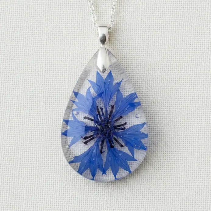 A clear, teardrop-shaped resin pendant on a silver chain, encapsulating a single small blue pressed flower.