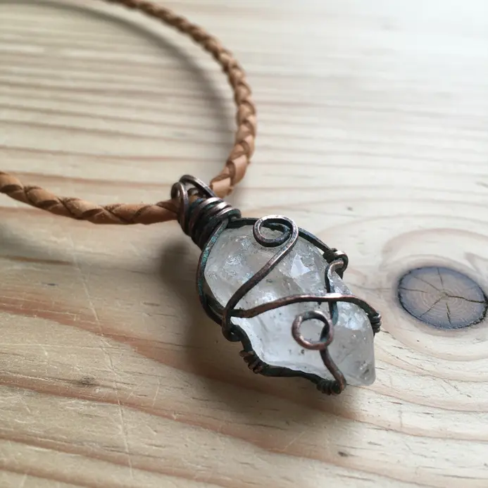 A clear, pointed raw quartz crystal wrapped at its base with oxidized copper wire on a leather cord.