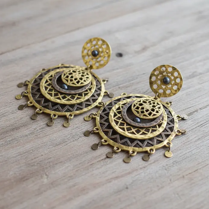 A pair of circular, hammered brass mandala earrings with detailed cut-out patterns and dangling bead accents.