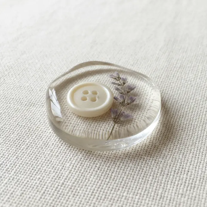 A round, clear resin pendant containing a dried lavender sprig and a single pearl button at its center.