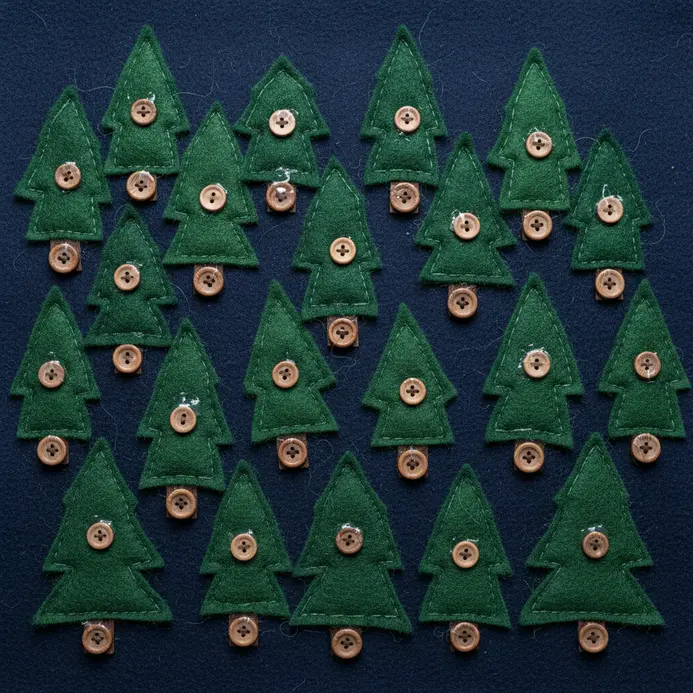A textile art piece with green felt pine trees against a blue felt sky, with brown buttons as tree trunks and yellow buttons as stars.
