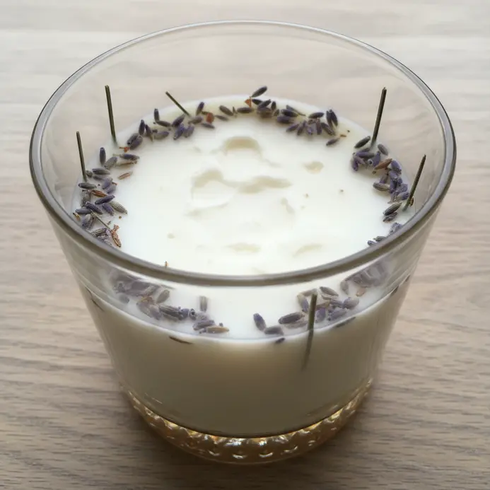 A clear glass jar containing a creamy soy candle with pressed lavender buds visible inside.