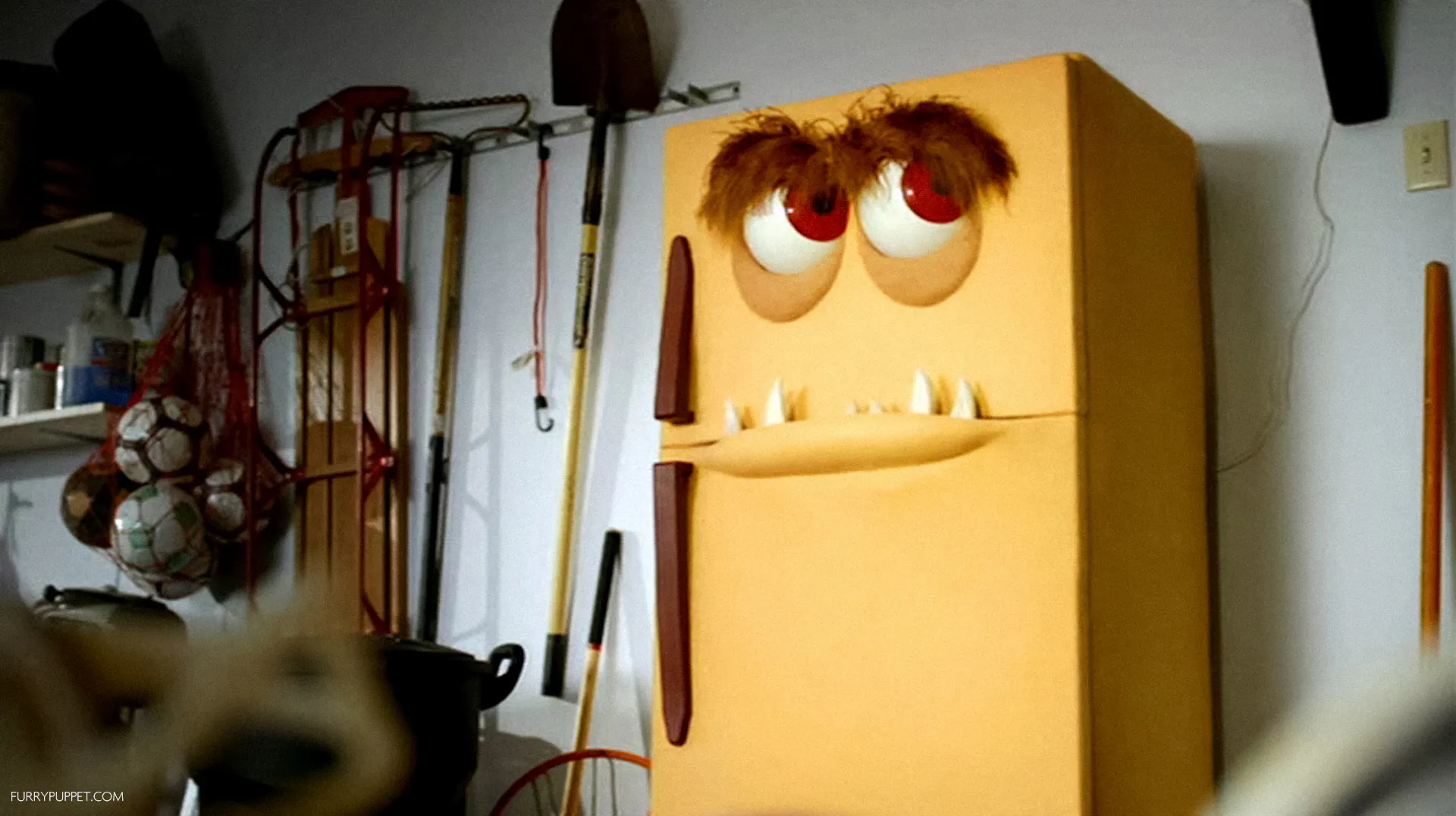 A yellow refrigerator in a garage transformed into a monster character with furry eyebrows, googly red eyes, and pointed teeth around the door seam.