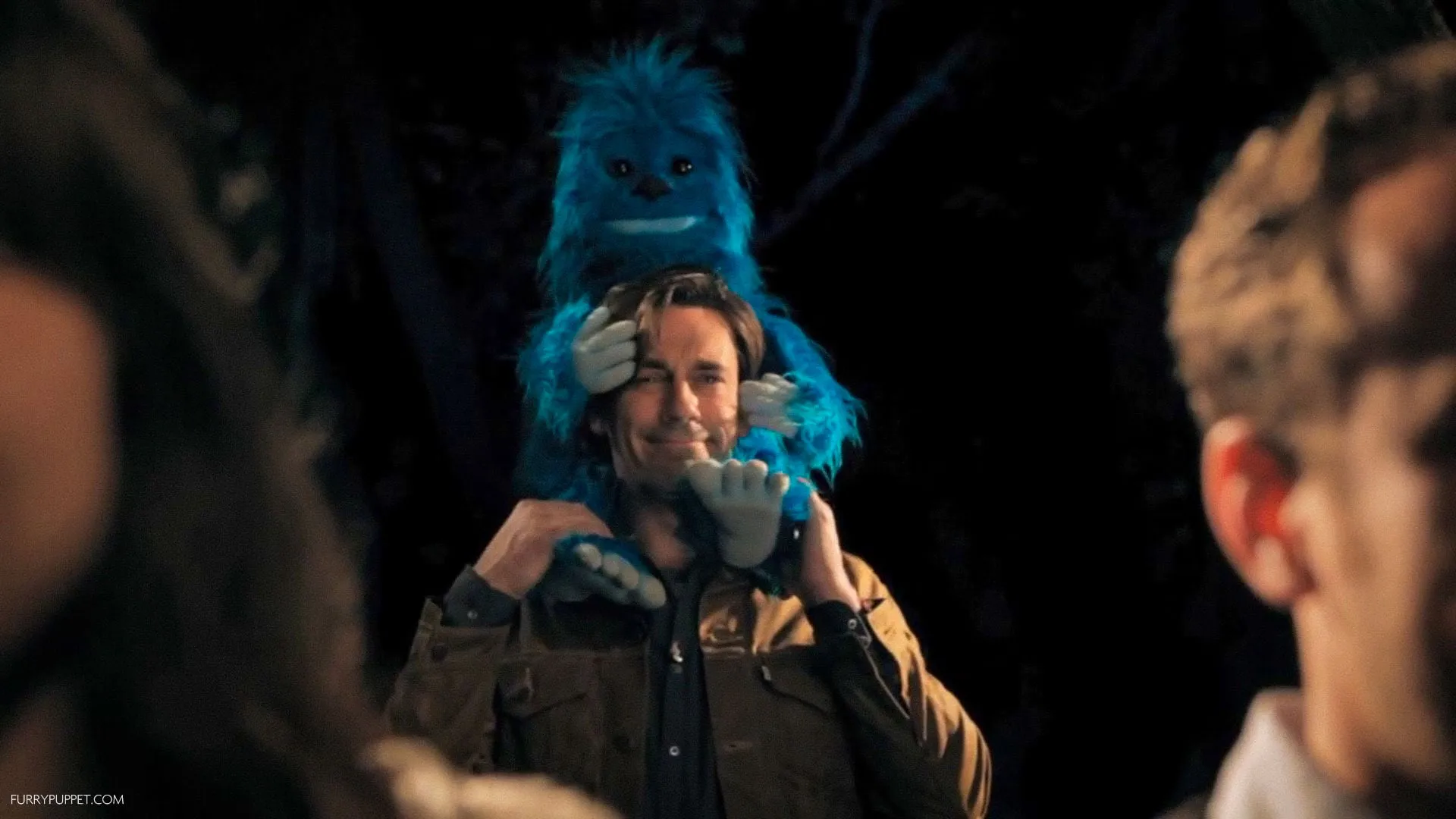A bright blue furry yeti puppet riding on Jon Hamm's shoulders with its hands covering his eyes at night.
