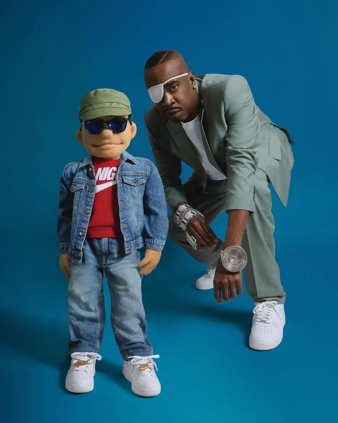 Slick Rick crouching beside a small puppet wearing an eye patch, denim jacket, and Nike sneakers against a blue backdrop.