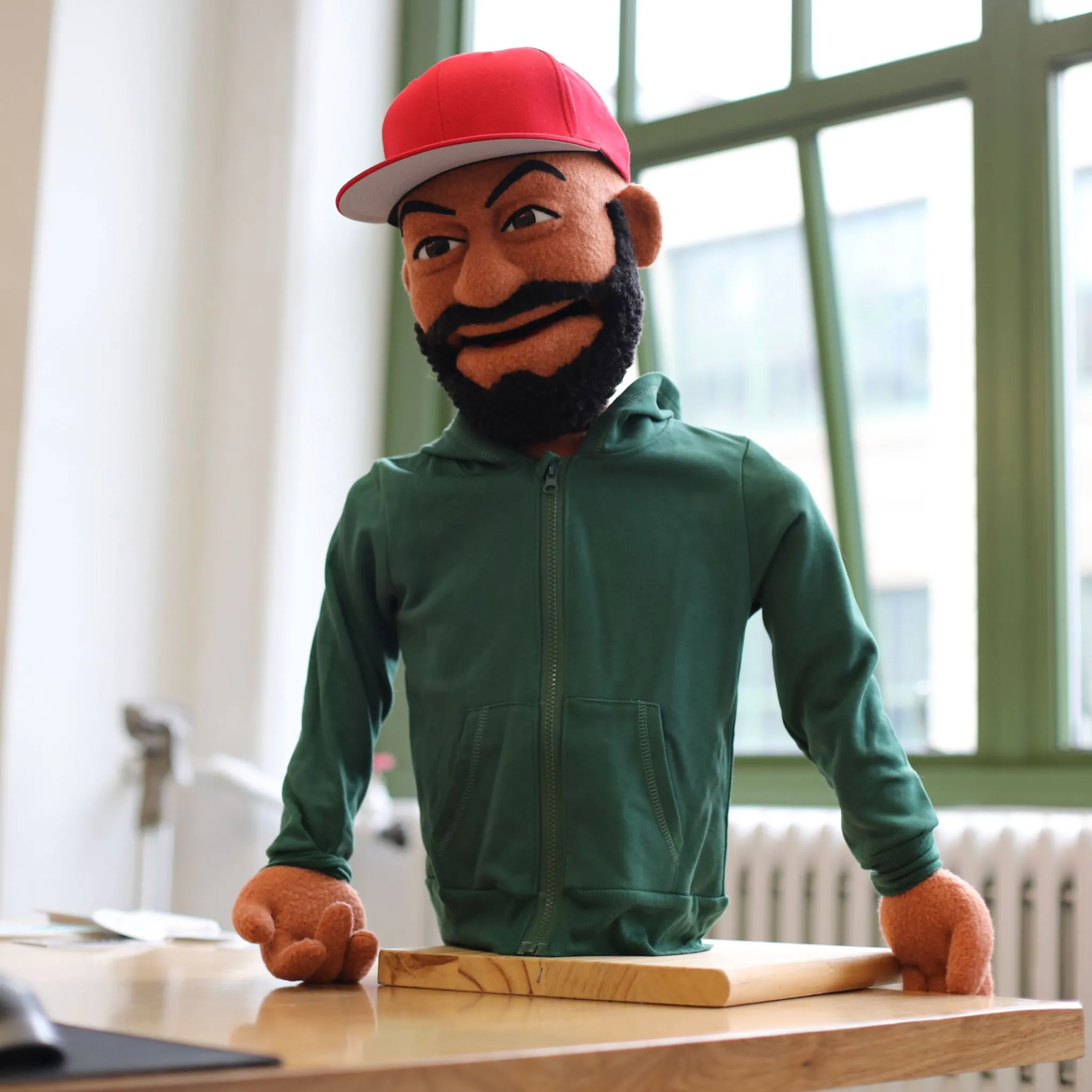 A bust puppet with a felted beard, red snapback cap, and green hoodie resting its arms on a wooden tabletop.