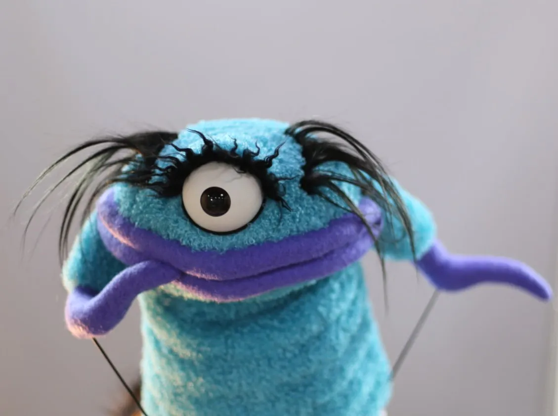 Close-up of a teal furry monster puppet with one large round eye, wild black brow hairs, and a purple mouth held open by visible control rods.