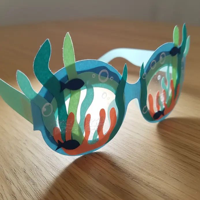 Blue paper glasses with small paper cutouts of fish, seahorses, and seaweed attached.