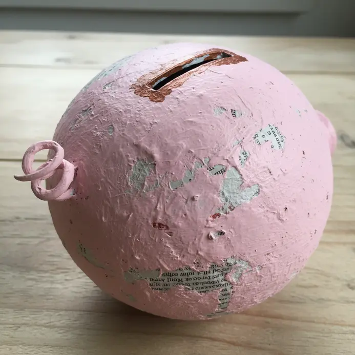 A pink paper mache piggy bank sculpture made from a balloon base, decorated with a curly tail and a coin slot on top.