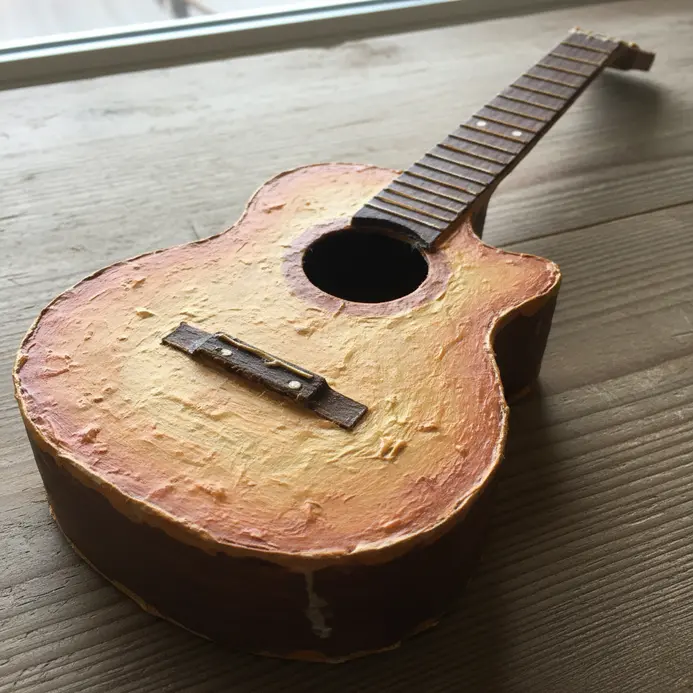 A brown paper mache acoustic guitar replica with detailed soundhole and painted strings, built on a cardboard base.