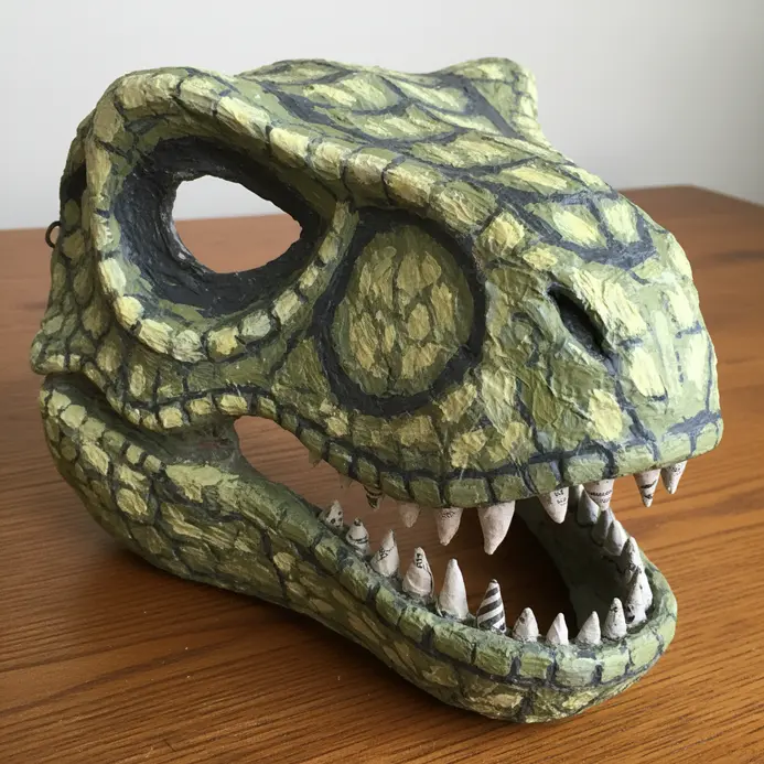 A green paper mache dinosaur mask with scales and teeth, formed over a balloon and held with an elastic strap.