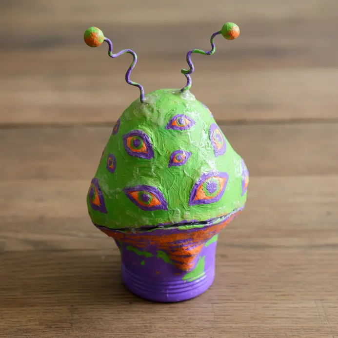 A green paper mache alien sculpture with multiple eyes and pipe cleaner antennae, built around a plastic bottle base.