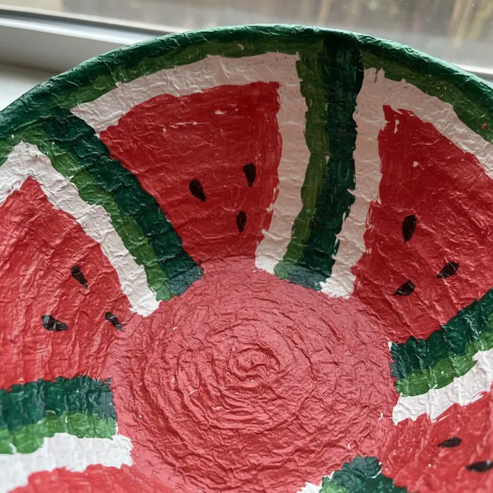 A wide, shallow paper mache bowl painted with a watermelon slice pattern, formed over an inflated balloon mold.