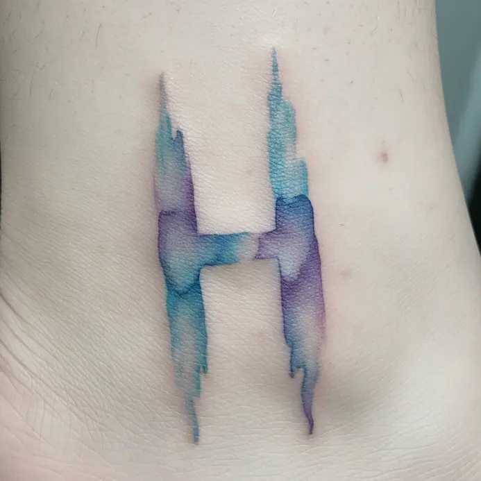 A letter H tattoo that mimics a brushstroke, with soft edges and watercolor-style blue and purple washes near the ankle bone.
