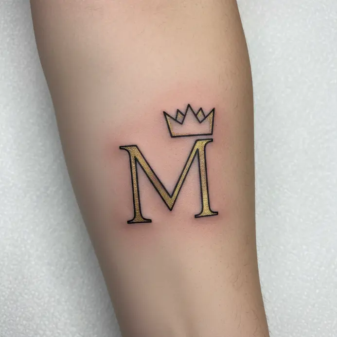 A very small, simple letter M tattoo with a tiny crown drawn above it, placed behind the earlobe.