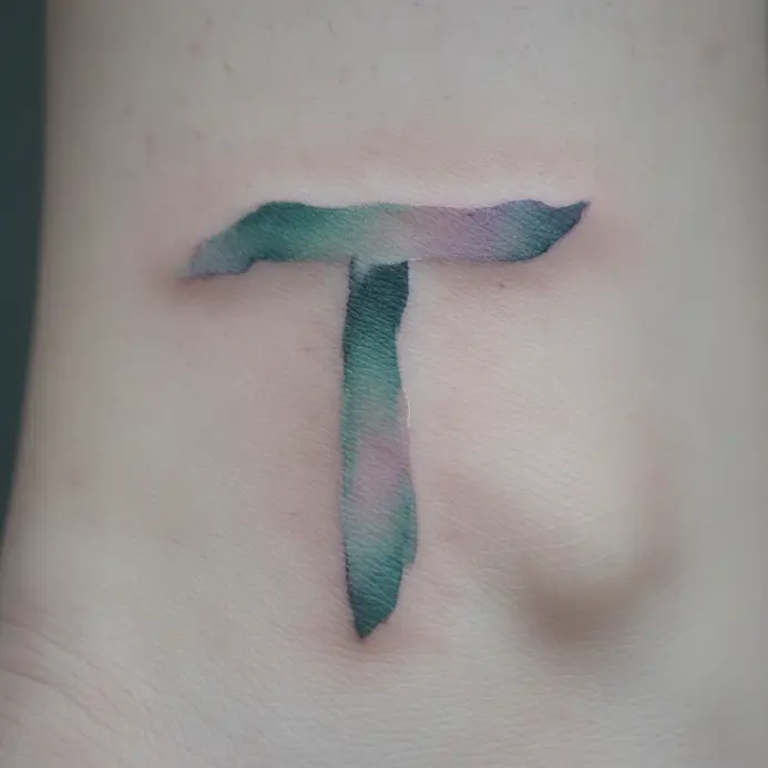 A letter T tattoo with soft, blended watercolor edges resembling a brushstroke on the ankle.