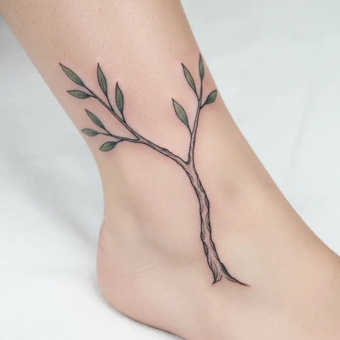 A black linework tattoo where the letter Y is formed by a slender tree branch with small leaves, wrapping an ankle.