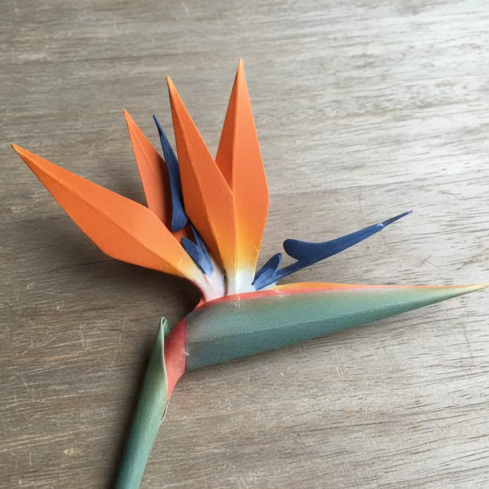 A modern table setting featuring one tall, orange and blue paper bird of paradise flower in a simple white vase.