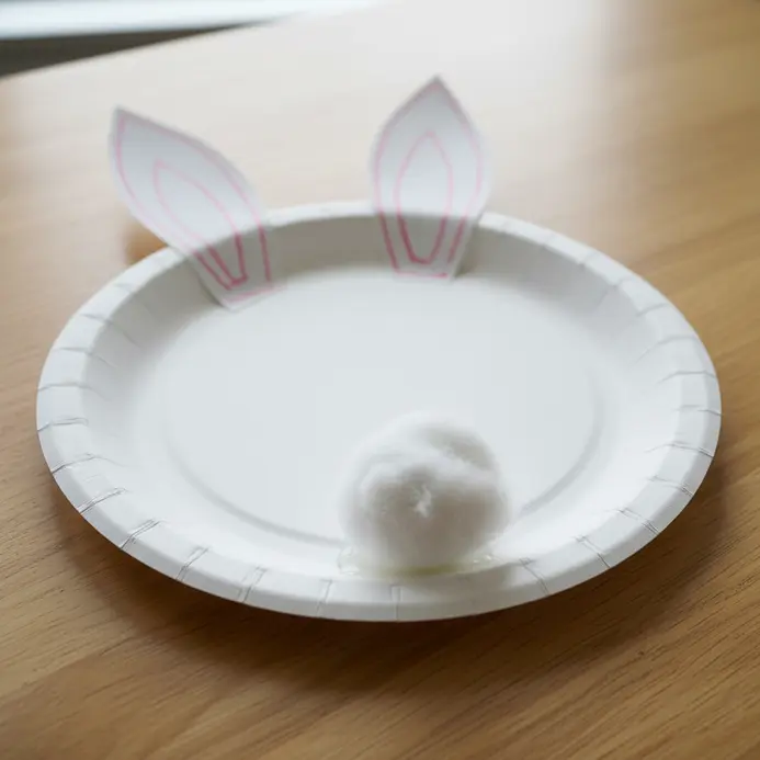 A white paper plate rabbit face with long pink-lined ears and a small cotton ball tail.