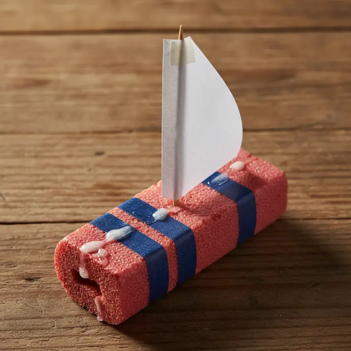 A red pool noodle boat with a striped paper sail floats in a kiddie pool.