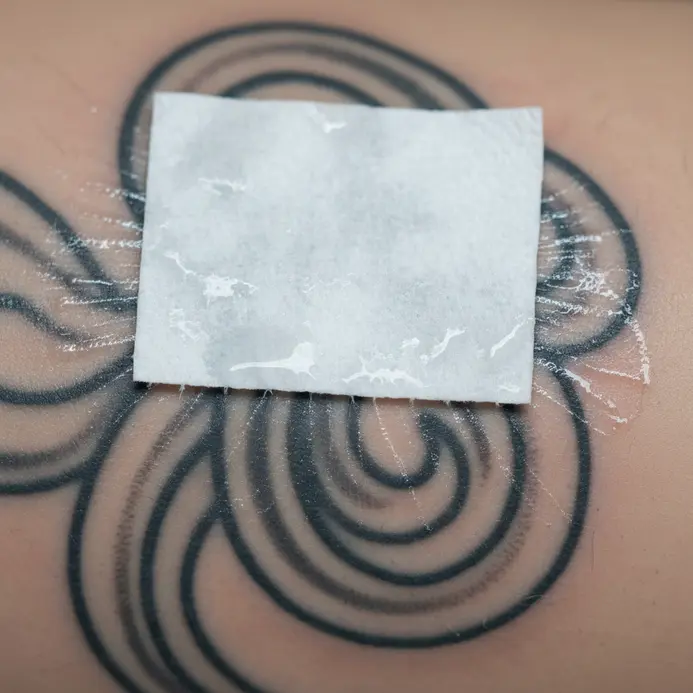 A single-use paper towel patting a freshly washed and wet tattoo dry on clean skin.