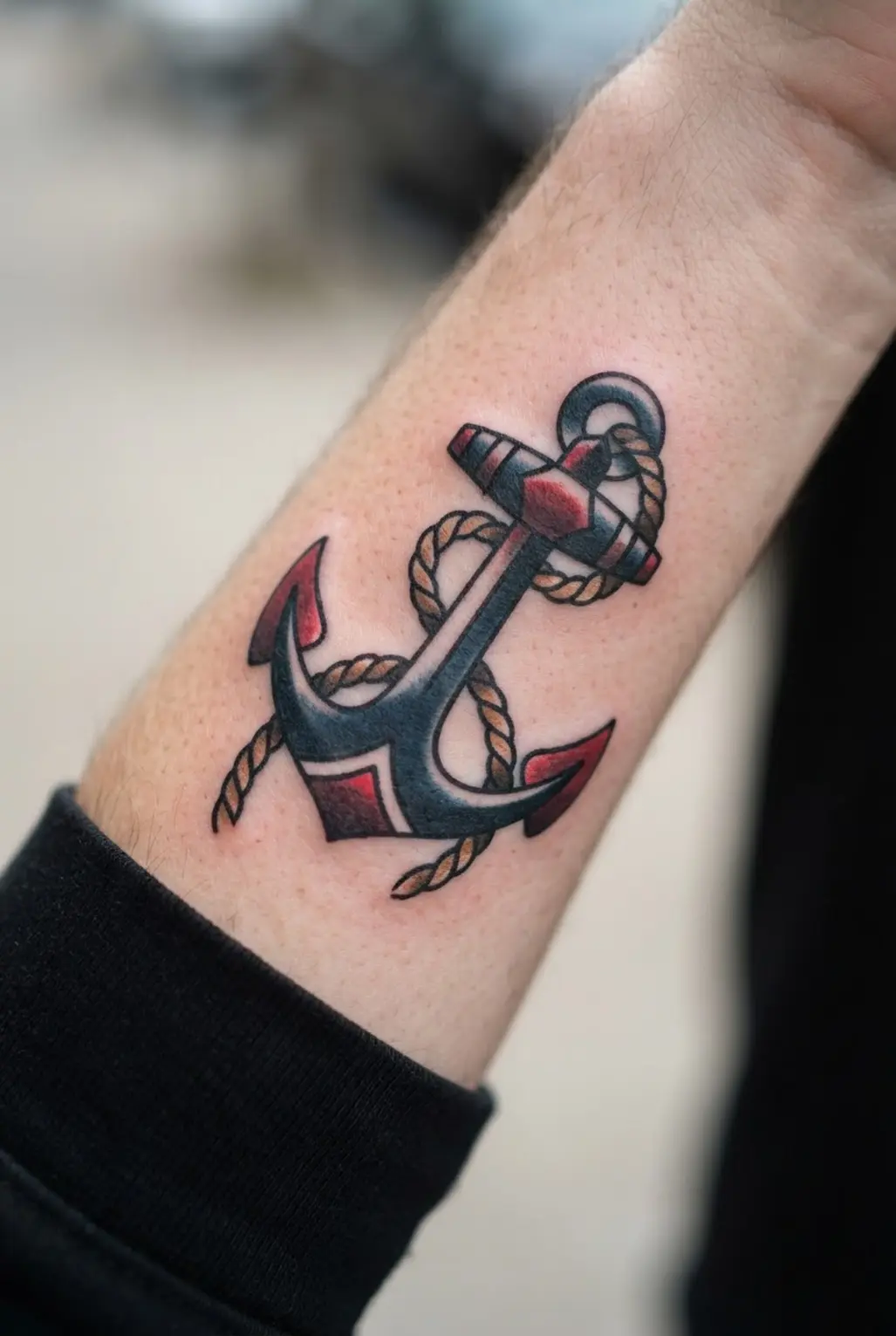 Anchor and rope forearm tattoo with navy blue shading, red accents, and a compact flat composition.