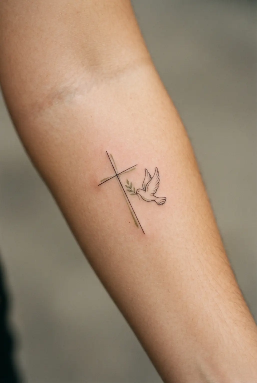 A small dove and olive branch sit beside a vertical cross on the forearm in soft black illustrative linework.