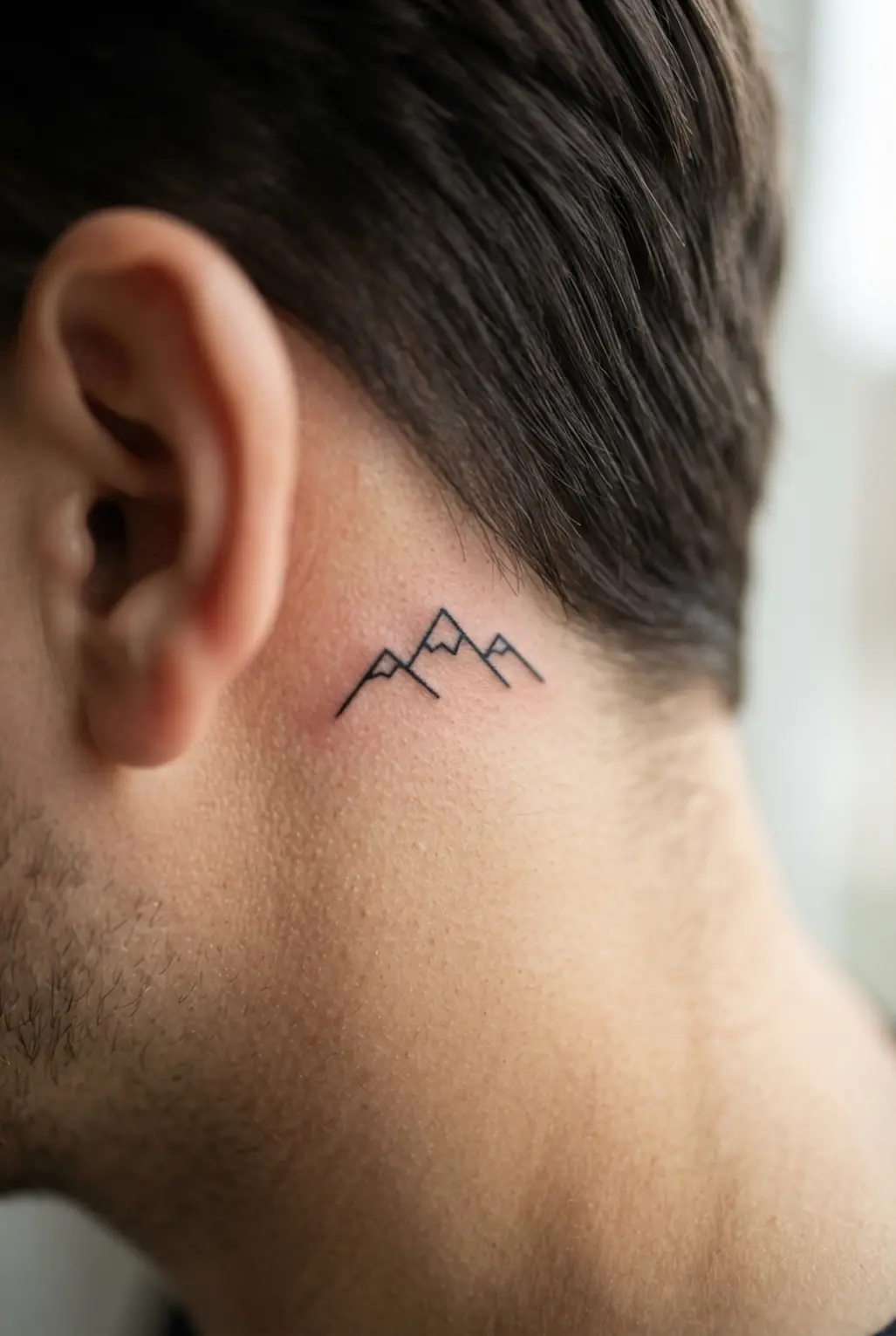 Fine-line black mountain range tattoo behind the ear, showing three clean peaks and a small masculine profile.