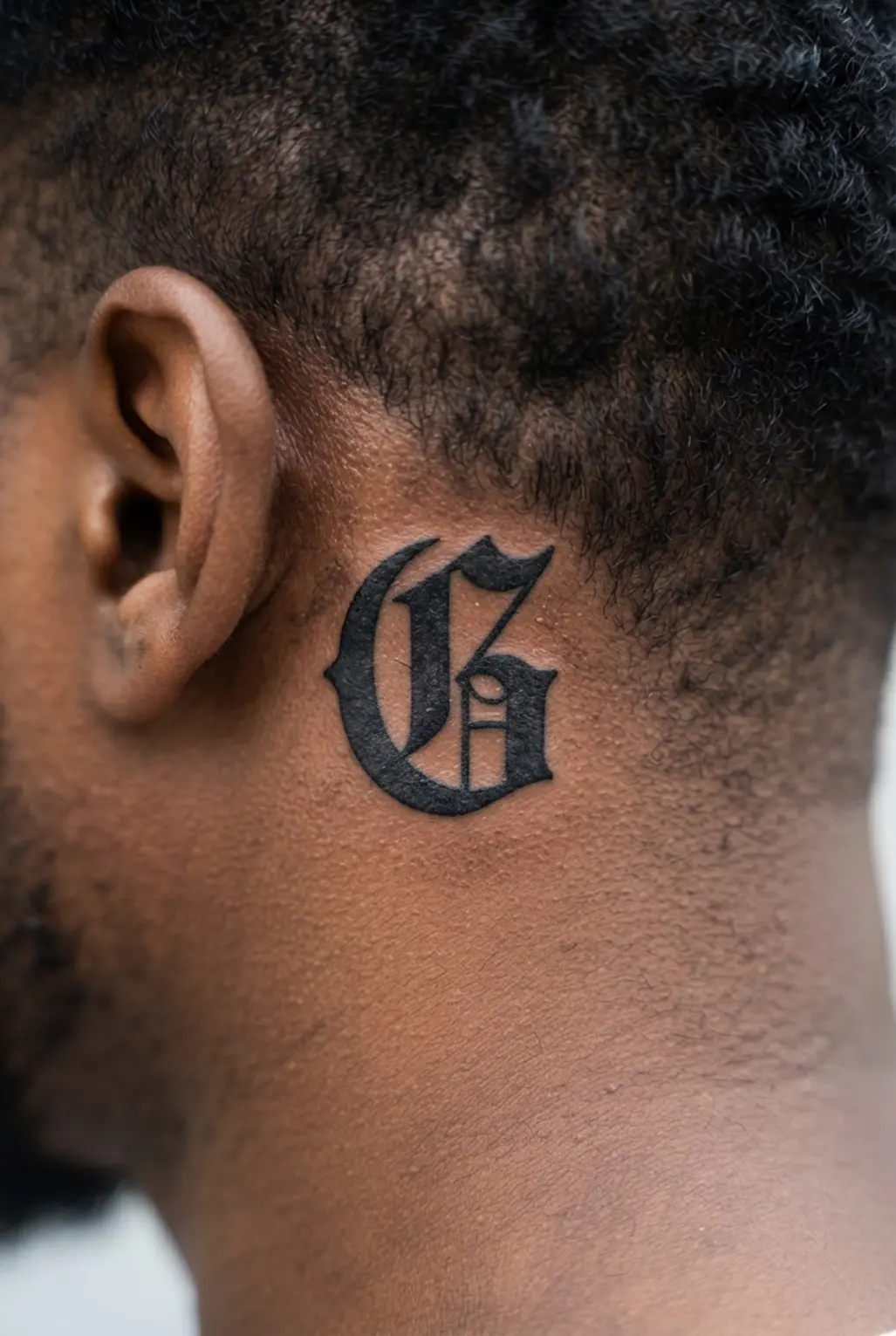 Classic Old English initial letter tattoo behind the ear, with heavy black strokes and pointed decorative serifs.