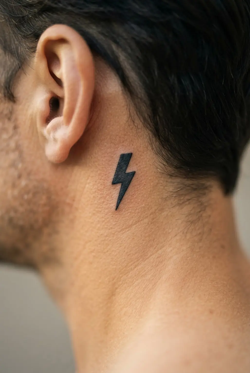 Simple black lightning bolt tattoo behind the ear, with a compact zigzag silhouette and sharp pointed ends.